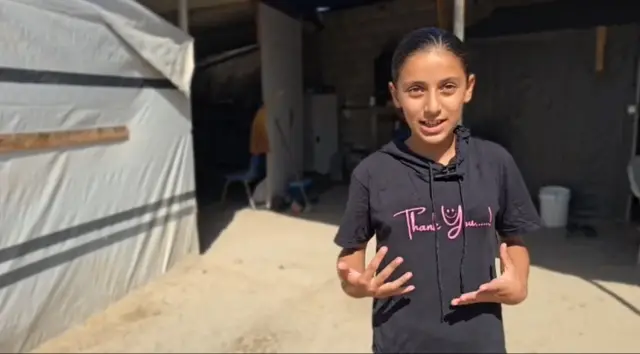 Sara stands in front of a building speaking while gesturing with her hands. She is wearing a black short-sleeved hoodie with the words "Thank You" on it.