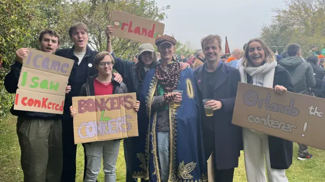 Orlando is stood in the middle wearing a navy blue cape. People are stood next to him and holding signs standing in front of trees.