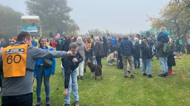 People were standing on the left practising playing conkers. People are crowded around an arena.