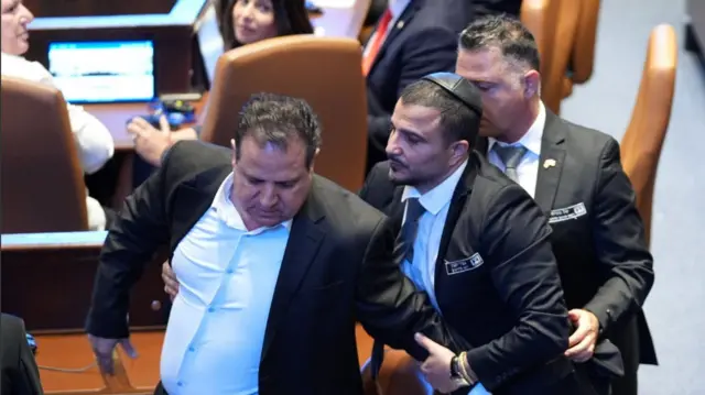 A member of Knesset is grabbed and escorted out by two members of security in black suit after disrupting Trump's speech in the Knesset