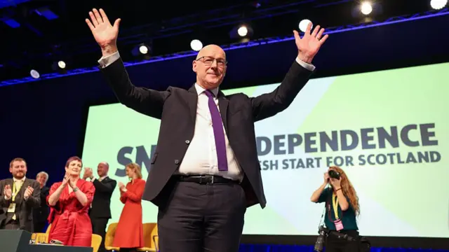 John Swinney in a suit with purple tie, he is bald and wears glasses. He is smiling and his hands are raised above his head in celebration. Behind him people clap and a woman with red hair uses a professional camera to take a picture. Sign behind them says SNP Independence, fresh start for Scotland.