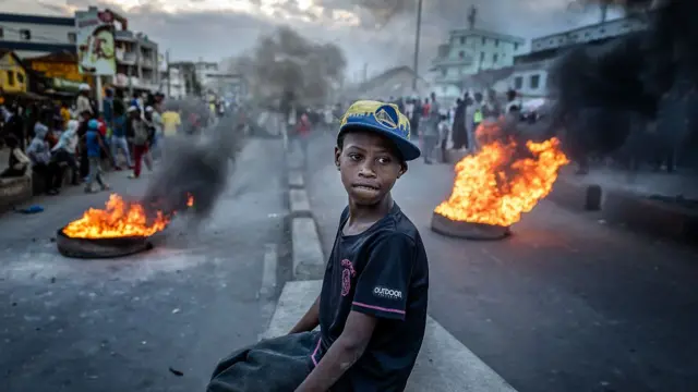 A boy sits atop a wall as two tyres burn behind him
