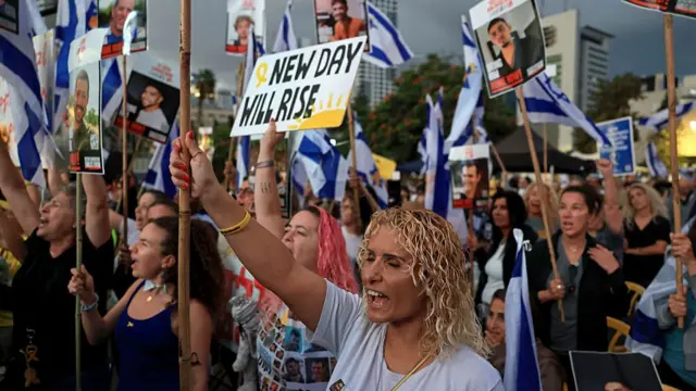Crowd of people standing, many holding placards with the photos of Israeli hostages. One sign reads 'New day will rise'.