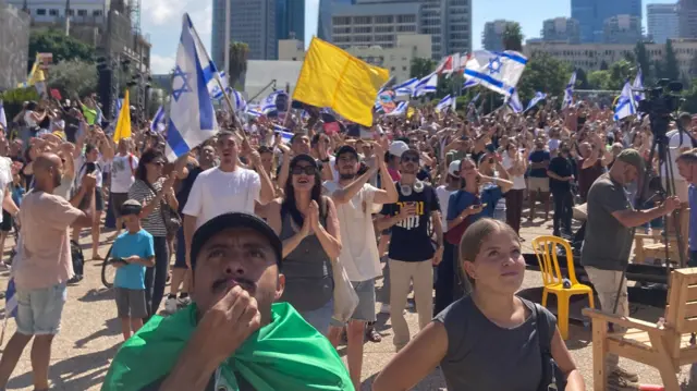 A crowd of people - some holding Israel flags and yellow flags, look to the sky and celebrate