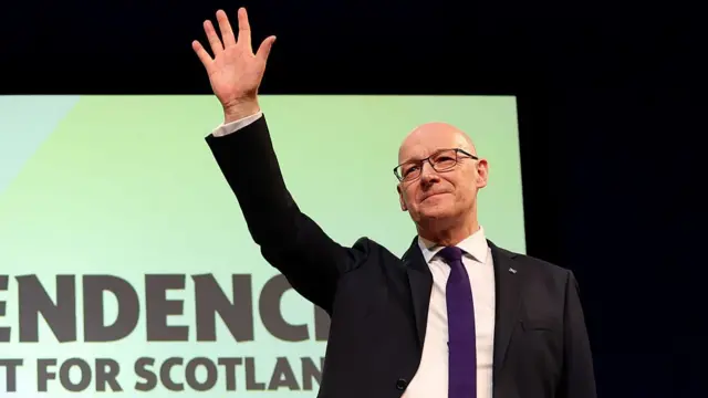 John Swinney, who is bald and wears glasses, raises his hand above his head to wave at crowd who are not in the image. He wears a black suit with purple tie.