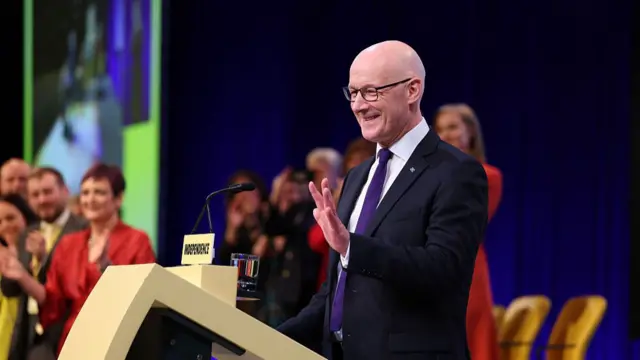 John Swinney stood behind podium waving.