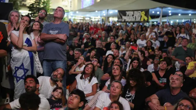 Large group of people, some seated on the floor and some standing, all looking up towards a screen (which can't be seen)