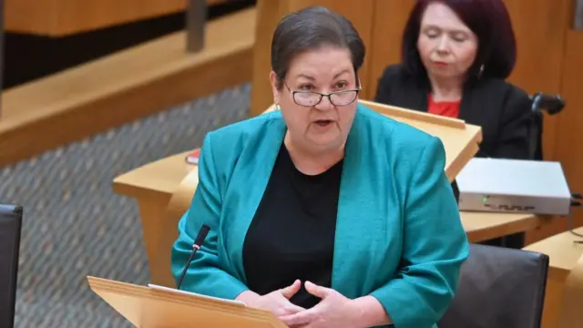 Jackie Baillie in a blue blazer and black t-shirt speaking behind a podium.
