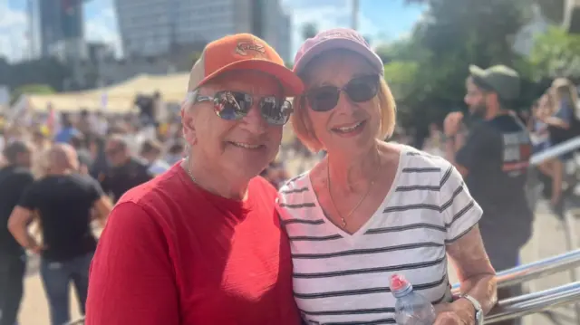 Harvey Betsalel and his wife smiling in Hostages Square, Tel Aviv. Both wear sunglasses and sunhats. Crowds and buildings in background.
