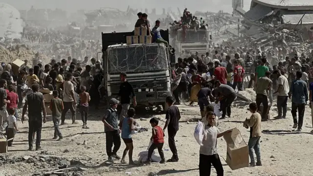 a thick crowd of Palestinians crowd around an aid druck with the devastation wround by Israel's bombardments in the background