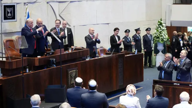President Donald Trump standing with Speaker Amir Ohana and Israel's President Isaac Herzog in the Knesset. Many politicians are standing and clapping while Trump points at someone off camera
