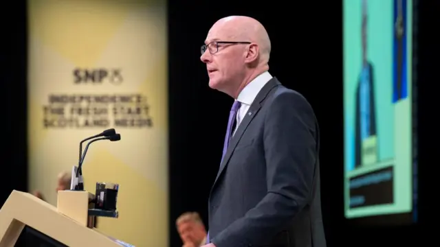 SNP leader John Swinney in a dark suit with purple tie speaking into a microphone behind a podium. The picture shows his profile as he is side on to the camera. Behind him is a large yellow banner that says SNP. People sit behind him and another person is stood on stage - both blurred.