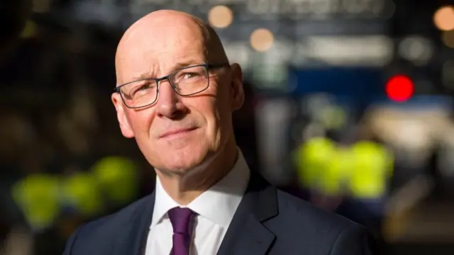 Head and shoulders shot of John Swinney, a bald man with rectangular glasses, who is wearing a navy blue blazer, a white shirt and a purple tie
