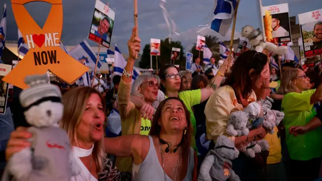 Crowd of people standing, many holding placards with the photos of Israeli hostages, other holding teddy bears. The sky shows the day breaking as the sun rises.