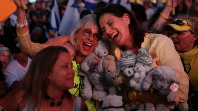 Two women hugging each other, both with big smiles, eyes closed, both holding several teddy bears. Crowds behind them holding the Israeli flag.