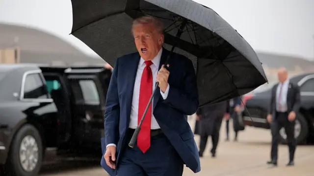 Donald Trump with his mouth open and holding an umbrella on an airport tarmac with secret service agents and vehicles in the background