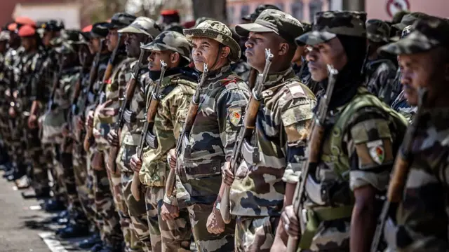 Armed soldiers in military fatigues stand in a horizontal line