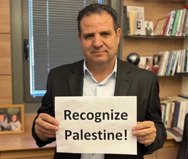 Ayman Odeh in home office holding sign which says 'Recognize Palestine'. He wears shirt and suit jacket.