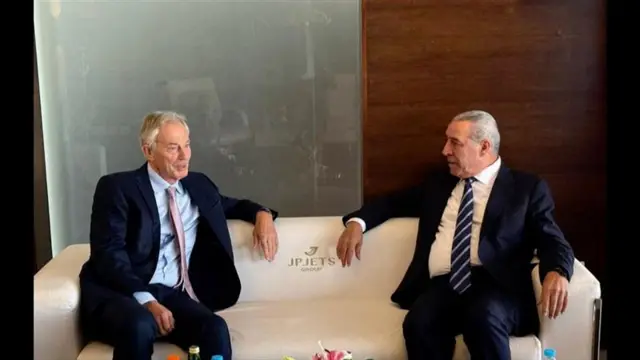 Two men sitting, both in dark suits, sitting at opposite ends of a cream couch, facing each other. One is smiling.