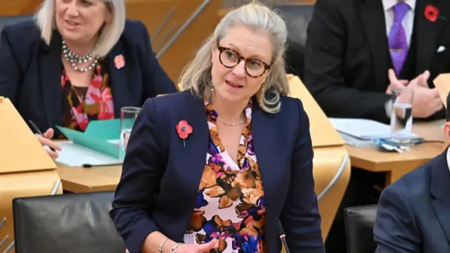 Rachael Hamilton with blonde hair and glasses wearing a floral top and blue blazer.