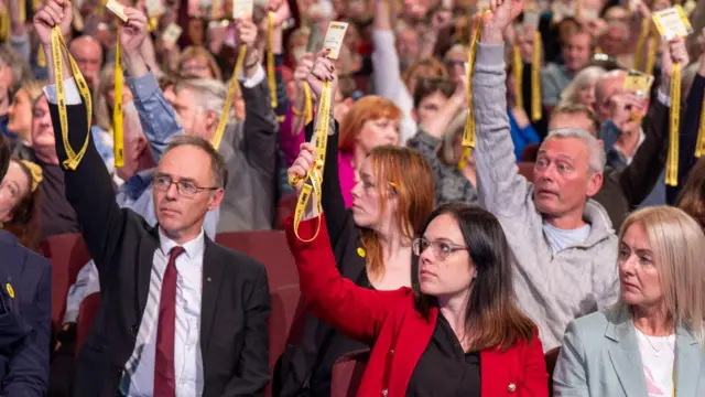 SNP members voting on independence strategy. Kate Forbes is in the middle in a red jacket holding a yellow pass.