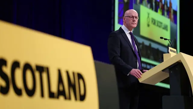 John Swinney standing behind podium that says Scotland