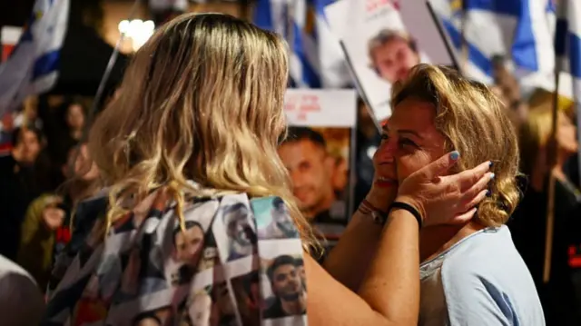 A woman with shoulder-length blonde hair, her back to the camera holds the cheeks of another woman as they wait for the release of Israeli hostages by Hamas