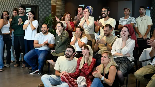 A group of people stare at a screen, they hold hands and many are crying
