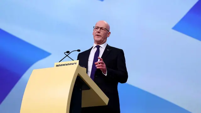 John Swinney, a bald man with glasses, stood behind a podium. Behind him is a Scottish flag.