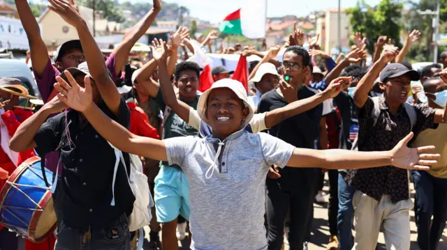 A group of protesters raise their arms. One carries a drum and a whistle can be seen also.