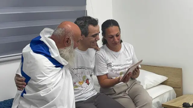 Three people sat on bed, embracing whilst looking at tablet screen