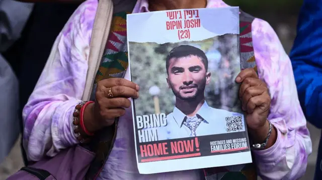 A person in a pink shirt holds up a photo of hostage Bipin Joshi with the words 'Bring him home now' on it.