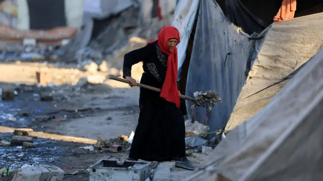 A Palestinian woman wearing a red head scarf cleans an area next to tents