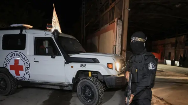 A Red Cross vehicle passes through a gate while a person with their face covered holds a gun