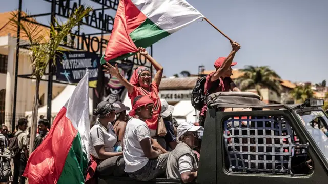 Protesters are piled in the back of a pick-up truck. One waves the flag of Madagascar.