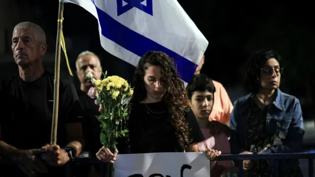 Woman standing wearing black, looking folorn, and leaning on railing in front of her. She's holding up a sign in Hebrew and holding a large bunch of yellow flowers in her right hand. It's night time and there are a small number of people standing next to and behind her.