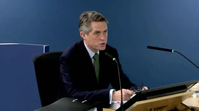Gavin Williamson in a black suit and green tie sits at a desk in a room with a microphone in front of him