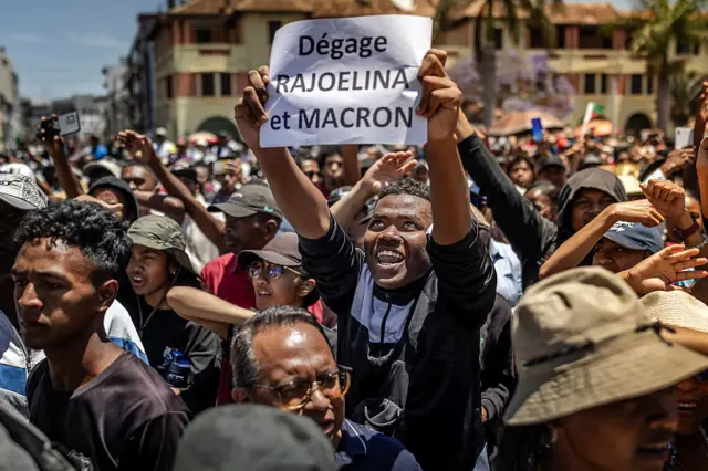 TOPSHOT - A protester holds a banner critical of Madagascar's President Andry Rajoelina and France's President Emmanuel Macron as residents gather for a civil society rally demanding Rajoelina's resignation in Antananarivo, on October 14, 2025. President Andry Rajoelina ratched up tensions in Madagascar on October 14, 2025 by dissolving the National Assembly to block a vote to force him out of office as thousands gathered for new protests calling for his resignation. Civil servants and trade unionists joined the large crowd in the capital in a fresh demonstration against Rajoelina after he made clear in an address from hiding late Monday that he did not intend to bow to demands that he quit. (Photo by Luis TATO / AFP) (Photo by LUIS TATO/AFP via Getty Images)