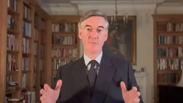 Jacob Rees-Mogg is sat in what looks like a study, with bookshelves behind him, a painting and a chandelier. He is wearing a black suit, stripey suit, and a black tie with white spots.