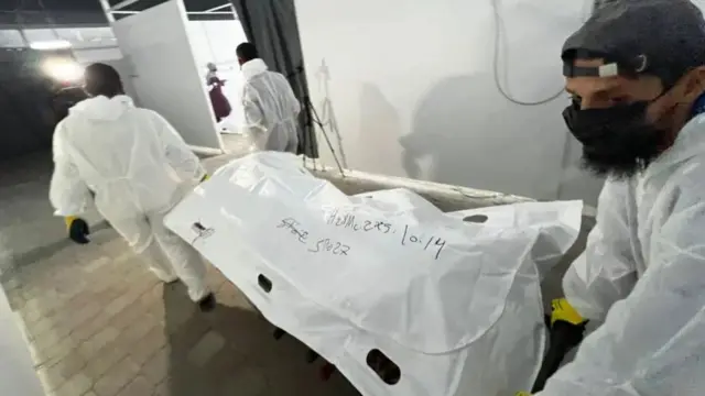 Three people roll a stretcher holding a white body bag, labelled with numbers