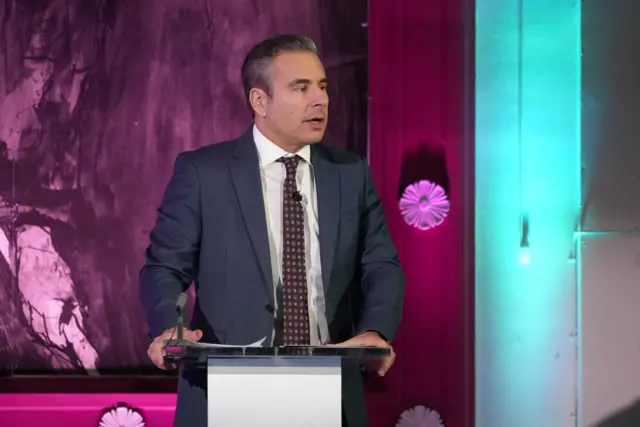 Nick Servini stood behind a podium in preparation for the by-election debate. He is wearing a suit with a burgundy tie. There are purple and blue lights in the background from the podium stage.