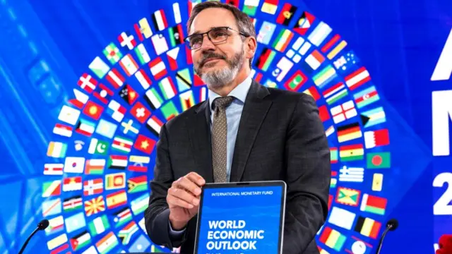 Man holds up a tablet with the words "World Economic Outlook" on it, while standing behind a circle made up of the flags of the world