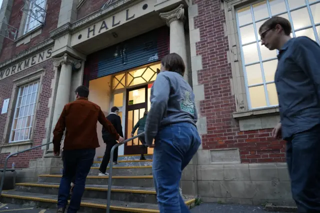 Several people walking in a row as they enter the Bedwas Workmen's Hall. They are wearing smart casual clothing and a light can be seen at the entrance.