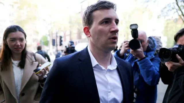 A headshot of Christopher Cash as he walks past the camera. Members of the press behind him take his photo