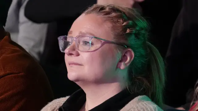 Jordan Morris yn gwisgo crys ddu a chardigan. Mae ei gwallt wedi clymu y'nôl ac mae ganddi sbectol.