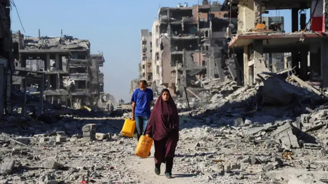 A man and woman carry water through destroyed buildings in Gaza City