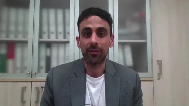 A man looks at the camera in a white tshirt and grey jacket