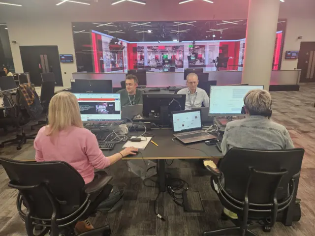 Four people sitting in chairs at a their desks. They are looking at computer monitors in a BBC office. The TV studio can be seen behind them.