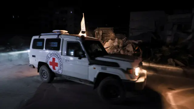 A white truck branded with the red cross logo casts its headlights on some rubble.