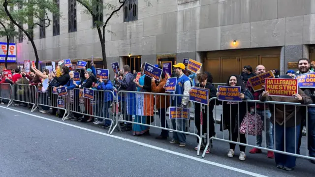 A group of people behind a fence holding campaign signs for Mamdani